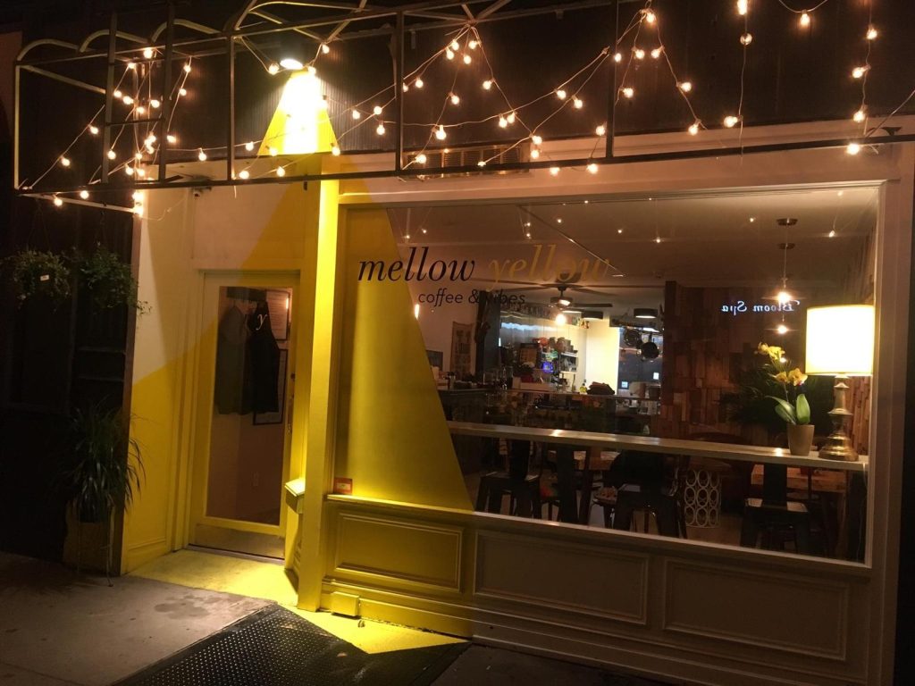 The exterior of Mellow Yellow, a cozy café in NYC, featuring warm decorative string lights, a bright yellow-painted entrance, and a welcoming ambiance with illuminated indoor seating visible through the large front window.