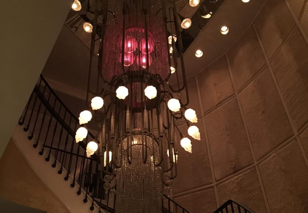 A grand, multi-tiered chandelier with cascading crystal strands and illuminated floral glass bulbs, hanging in a dimly lit staircase with a curved railing.